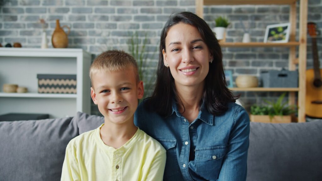 Foster With Us Luncheon Portrait of happy young family mother and little son smiling sitting together indoors looking at camera. Motherhood, childhood and apartment concept.